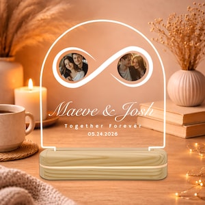 Peut inclure: Veilleuse LED personnalisée avec une base en bois. Le panneau en acrylique transparent présente un symbole infini avec deux encarts photo, les noms "Maeve & Josh", le texte "Together Forever" et la date 05.24.2026.