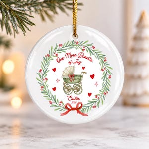 May include: A round white ceramic ornament featuring a green wreath with red berries. In the centre is a vintage-style green baby carriage, red hearts, and the text One More Bundle of Joy, Carter, and 2026. A red ribbon bow is at the bottom.