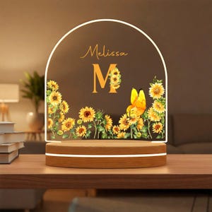May include: A personalized night light with a wooden base and an arched acrylic panel. The panel features the name "Melissa" and a large "M" decorated with sunflowers, along with a yellow butterfly. The light emits a warm glow.
