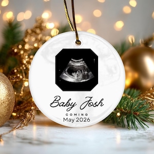 May include: A round white ceramic ornament featuring a black and white ultrasound image at the top. Below the image, the text reads Baby Josh in a black script font, followed by the text COMING May 2026 in a simple black sans-serif font.