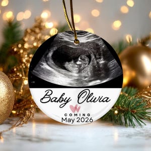 May include: A round ceramic ornament featuring a black and white ultrasound image of a fetus at the top. The bottom section is white with the black text Baby Olivia, two small pink hearts, and the black text COMING May 2026. It hangs by a gold cord.