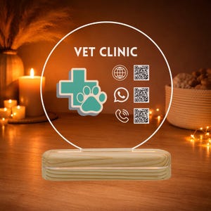 Op de afbeelding: Verlicht bord voor een dierenkliniek, met de tekst "VET CLINIC" en een turquoise kruis met een pootafdruk. Het bevat contactpictogrammen en QR-codes, op een houten voet. De achtergrond is zacht verlicht.