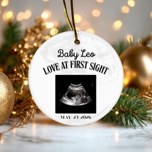 Può includere: Un ornamento rotondo in ceramica bianca con un'immagine ecografica in bianco e nero al centro. Il testo recita Baby Leo, LOVE AT FIRST SIGHT e MAY 23 2026. Un cordino dorato è fissato in alto per appendere questo oggetto decorativo.