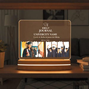 May include: A personalized graduation night light with a wooden base. The clear acrylic panel displays the text "EMILY JOURNAL, UNIVERSITY NAME" and the names Emily, Bella, Janet, and Olivia. Two photos of graduates are featured. The date "MAY 16, 2024" is also included.