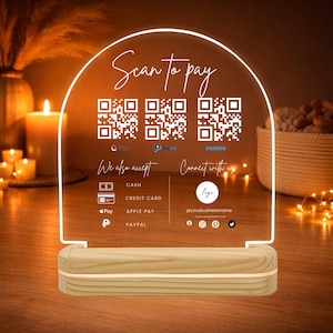 Op de afbeelding: Een helder acryl bord met een houten voet, verlicht met warm wit licht. Het bord leest "Scan to pay" met QR-codes voor G Pay, PayPal en Venmo. Het vermeldt ook geaccepteerde betaalmethoden: contant, creditcard, Apple Pay en PayPal.