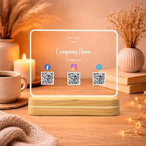 May include: A clear acrylic business card holder with a wooden base. The card displays the text "Your Logo Here" and "Company Name", along with social media icons and QR codes. The background includes a candle, coffee cup, and decorative plants.