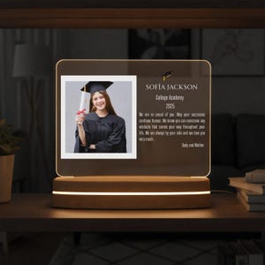 May include: A graduation photo display featuring a photo of a graduate in a cap and gown, with text that reads "SOFIA JACKSON College Academy 2025". The display is illuminated by a warm light and sits on a wooden base.