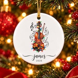 Könnte beinhalten: Ein rundes, weißes Keramikornament mit einer braunen Geige, die mit bunten Weihnachtslichtern und schwarzen Noten verziert ist. Unter der Geige steht der Name James in schwarzer Schreibschrift, gefolgt von der Jahreszahl 2026.