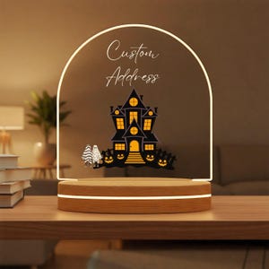 May include: An arched clear acrylic sign with a light-up wooden base. The sign features the text Custom Address in white script, a black haunted house illustration with glowing yellow windows, and two small ghost figures holding orange pumpkins.