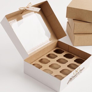 May include: A square cardboard box with a clear plastic window on the lid, featuring a twelve-compartment insert for baked goods. The exterior is white with brown interior sides and the text The Artisan's Oven Fine Baked Goods Est. 2021.