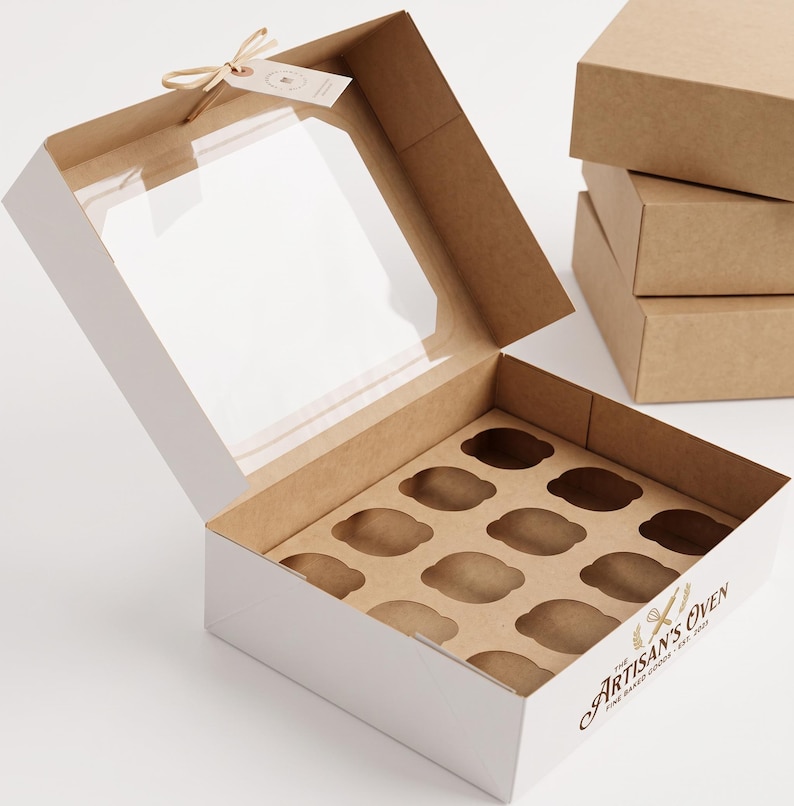 May include: A square cardboard box with a clear plastic window on the lid, featuring a twelve-compartment insert for baked goods. The exterior is white with brown interior sides and the text The Artisan's Oven Fine Baked Goods Est. 2021.