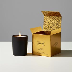 Könnte beinhalten: Eine brennende schwarze Kerze in einem Glas neben einer goldenen Schachtel. Die Schachtel ist geöffnet und zeigt ein Blumenmuster auf der Innenseite des Deckels. Auf der Schachtel stehen die Wörter "JAUNE" und "Winter Wonderland".