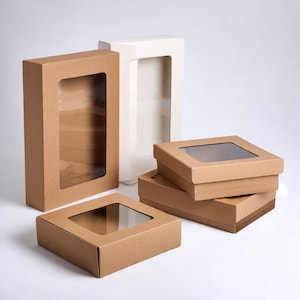 Pu&ograve; includere: Una collezione di scatole di cartone rettangolari e quadrate dotate di finestre in plastica trasparente sui coperchi. Il set include diverse scatole in carta kraft marrone e una scatola bianca alta, tutte con una struttura semplice e funzionale.