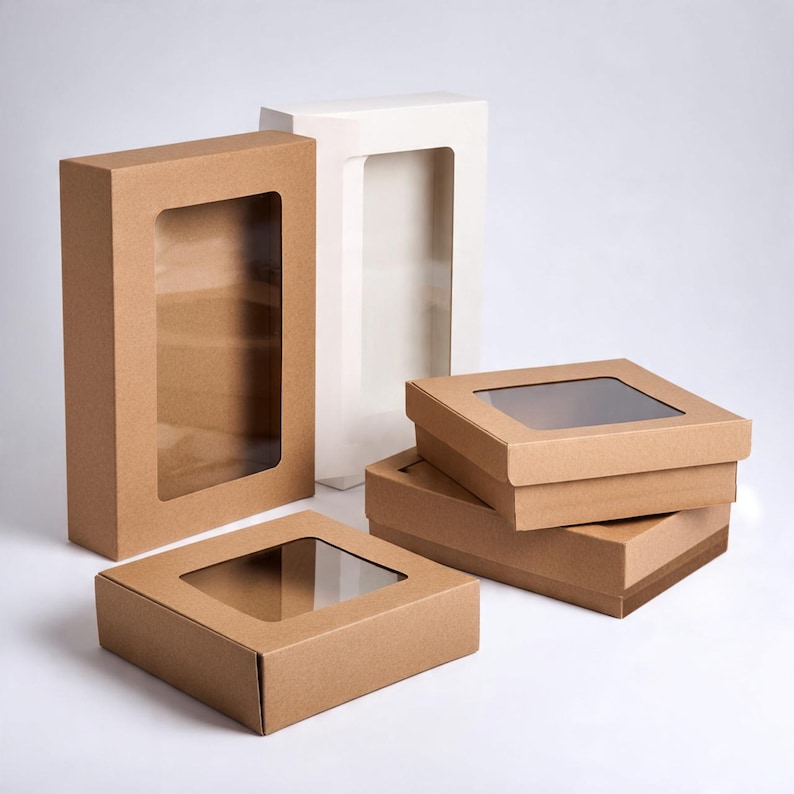 Pu&ograve; includere: Una collezione di scatole di cartone rettangolari e quadrate dotate di finestre in plastica trasparente sui coperchi. Il set include diverse scatole in carta kraft marrone e una scatola bianca alta, tutte con una struttura semplice e funzionale.