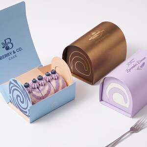 May include: Three rectangular cake boxes with curved tops. One light blue box is open, revealing a sliced cake roll topped with blueberries and purple cream. A bronze box features a Chef's Signature logo, and a lavender box is labeled Lavender Dream.