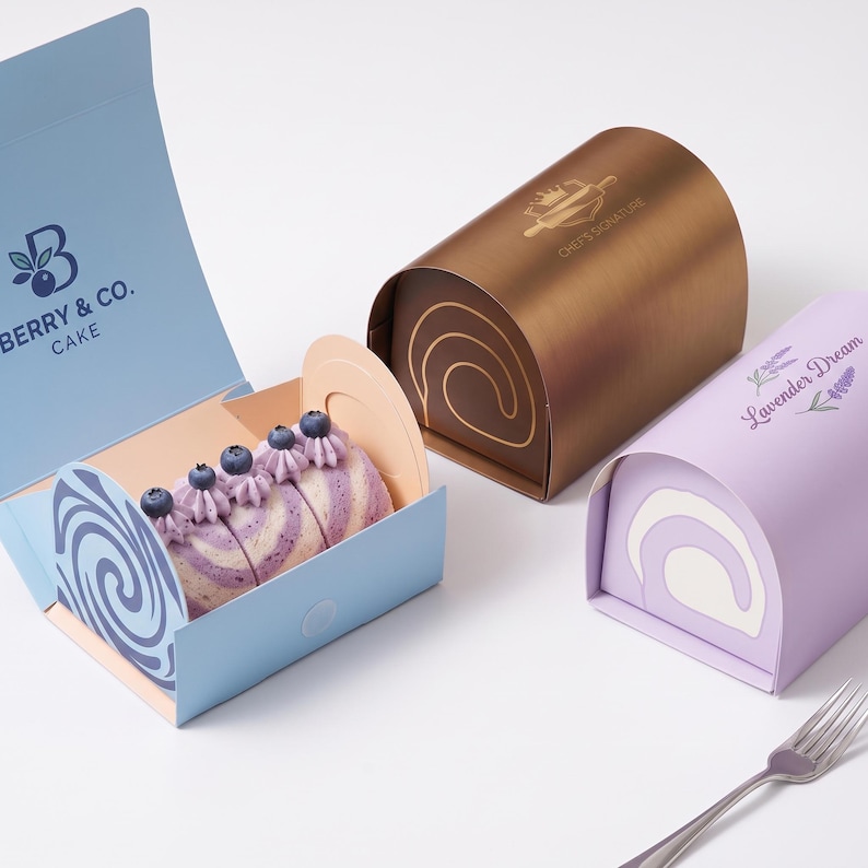 May include: Three rectangular cake boxes with curved tops. One light blue box is open, revealing a sliced cake roll topped with blueberries and purple cream. A bronze box features a Chef's Signature logo, and a lavender box is labeled Lavender Dream.