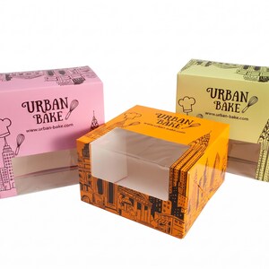 May include: Three rectangular cardboard boxes in pink, orange, and light yellow, each featuring a clear plastic window, black line art of city skylines, a chef hat, a whisk, and the text "URBAN BAKE" with the website "www.urban-bake.com".