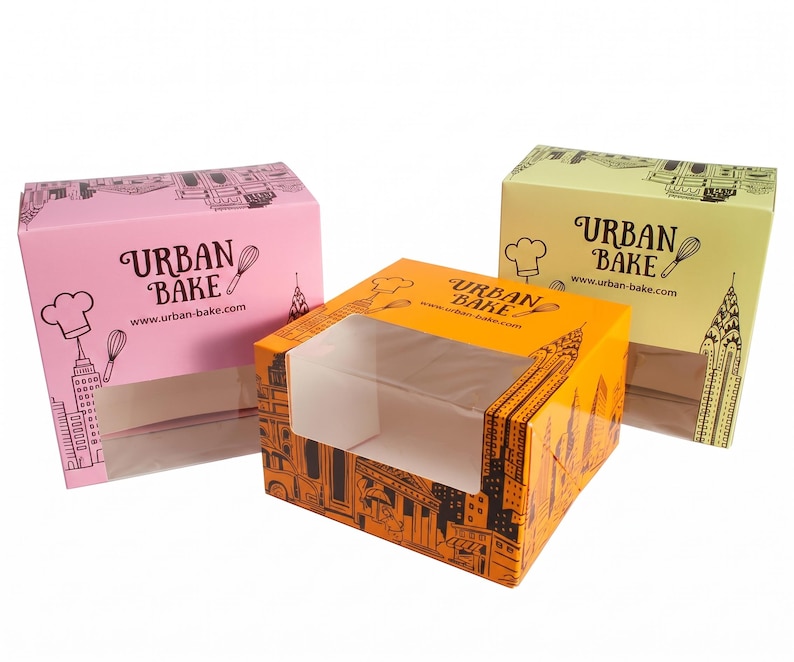 May include: Three rectangular cardboard boxes in pink, orange, and light yellow, each featuring a clear plastic window, black line art of city skylines, a chef hat, a whisk, and the text "URBAN BAKE" with the website "www.urban-bake.com".