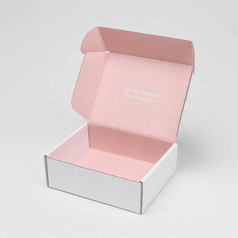 Pu&ograve; includere: Una scatola di spedizione rettangolare in cartone aperta con esterno bianco e interno rosa tenue. Il coperchio interno presenta il testo stampato in bianco Grace Eleyae e la frase Live confidently in un carattere sans-serif.