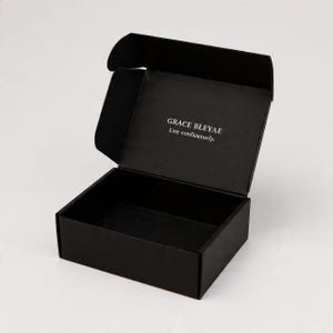 Pu&ograve; includere: Una scatola di spedizione rettangolare in cartone nero opaco con coperchio aperto. L'interno del coperchio presenta il testo stampato in bianco "GRACE BLEYAE" sopra la frase "Live confidently." La scatola &egrave; vuota e ha un design strutturato.