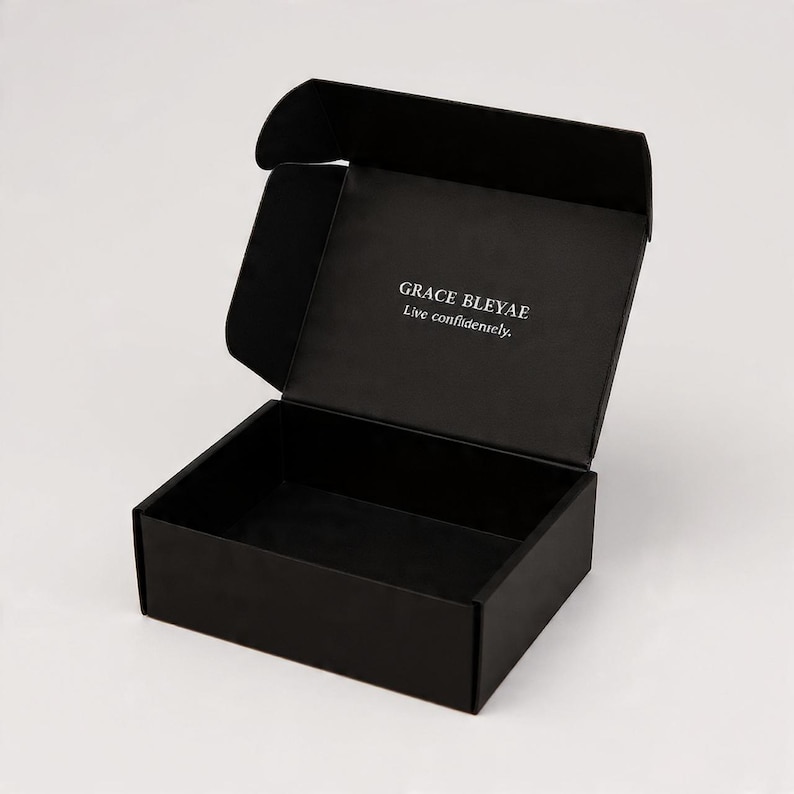 Pu&ograve; includere: Una scatola di spedizione rettangolare in cartone nero opaco con coperchio aperto. L'interno del coperchio presenta il testo stampato in bianco "GRACE BLEYAE" sopra la frase "Live confidently." La scatola &egrave; vuota e ha un design strutturato.