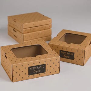 Puede incluir: Una pila de cuatro cajas de cartón marrón y dos cajas individuales con ventanas de plástico transparente. Cada caja presenta un patrón de cruces marrón oscuro y una etiqueta negra con el texto HOME BAKED Enjoy.