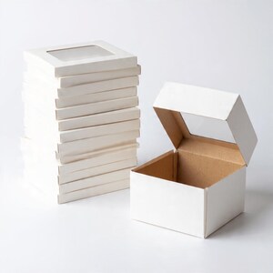 Pu&ograve; includere: Una pila di scatole quadrate in cartone bianco con finestre in plastica trasparente sui coperchi, accanto a una singola scatola in cartone bianco aperta con una finestra trasparente sul coperchio e una finitura interna marrone naturale.
