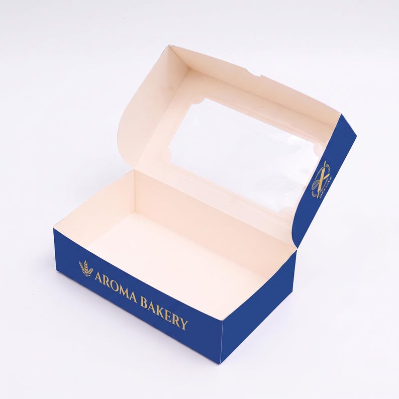 May include: A rectangular cardboard bakery box with a hinged lid featuring a clear plastic window. The exterior is deep blue with gold text reading AROMA BAKERY and a wheat stalk logo. The interior is a light cream color, providing a clean space.