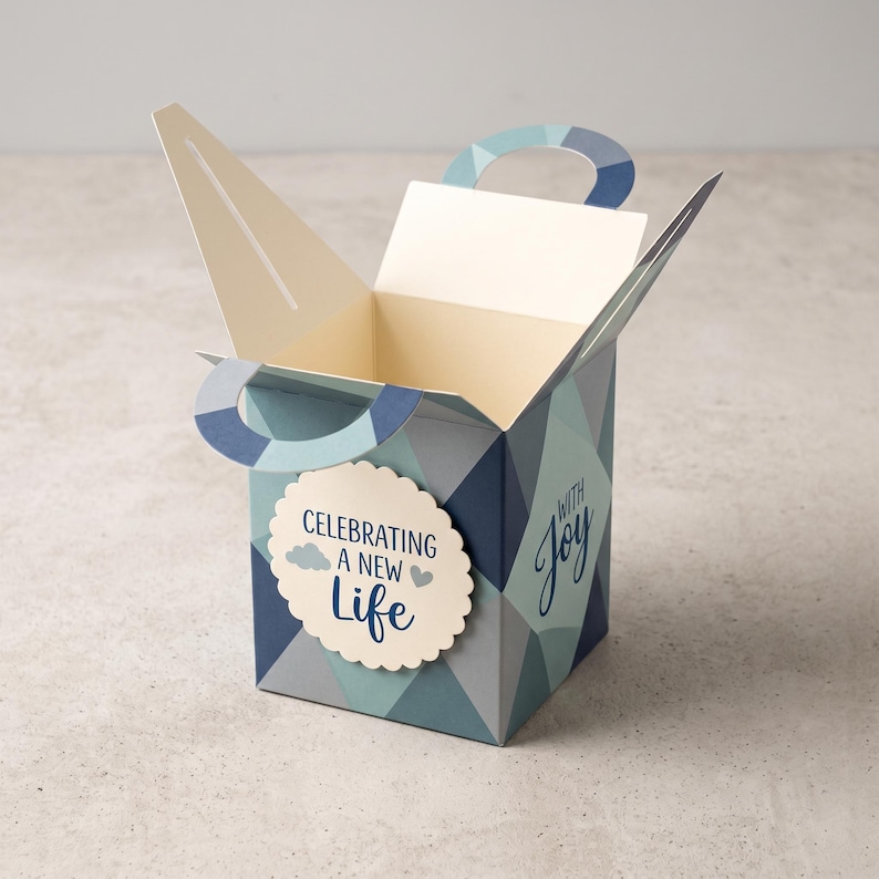 May include: A square cardboard gift box with a geometric pattern in shades of blue and gray. The box features two integrated handles and a scalloped circular tag on the front that reads "CELEBRATING A NEW LIFE" with "WITH JOY" printed on the side.