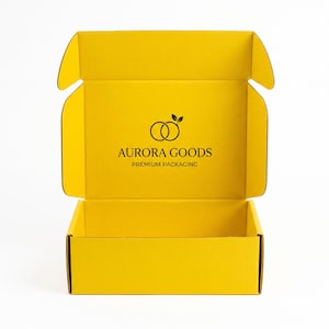 Op de afbeelding: Een felgele kartonnen verzenddoos met een open deksel. Het binnenste achterpaneel is voorzien van een zwart logo bestaande uit twee in elkaar grijpende cirkels met twee blaadjes erboven, en de tekst AURORA GOODS PREMIUM PACKAGING in het zwart.