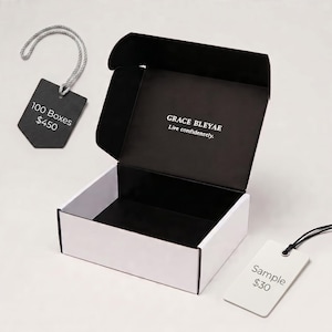 Pu&ograve; includere: Una scatola di spedizione rettangolare in cartone aperta con esterno bianco e interno nero. L'interno del coperchio presenta il testo bianco Grace Bleyae Live confidently. Sono presenti due etichette rettangolari con cordini, una nera e una bianca.