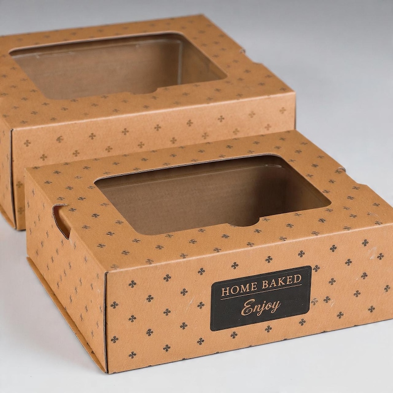 May include: Two rectangular brown cardboard boxes with clear plastic windows on top. The boxes feature a repeating pattern of small black four-pointed symbols. A black rectangular label on the front reads HOME BAKED Enjoy in white text.