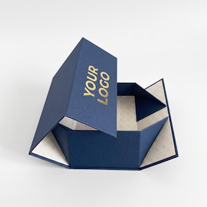 Könnte beinhalten: Eine zusammenklappbare marineblaue Geschenkbox mit Magnetverschluss. Auf dem Außendeckel befindet sich der goldfarbene Schriftzug YOUR LOGO. Das Innere ist mit cremefarbenem Papier ausgekleidet, das ein dezentes, geometrisches Punktmuster aufweist.