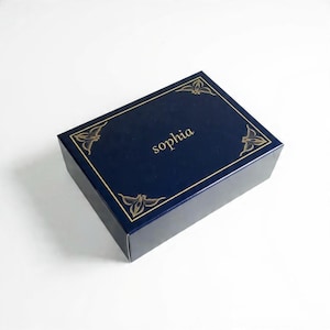 May include: A rectangular navy blue gift box featuring a gold-colored border and decorative corner accents. The name "sophia" is printed in gold-colored lowercase letters in the center of the lid. The box has a smooth, matte finish.