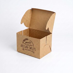 May include: A light brown cardboard box with an open lid. The front panel features a dark brown printed logo with the text Artisan Sweets Bakery, accompanied by illustrations of a wheat stalk, a whisk, a rolling pin, a cupcake, and a croissant.