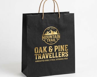 May include: A black paper shopping bag with two brown twisted paper handles. The front features a gold logo with the text MOUNTAIN TRAIL CO. EST. 2018, followed by the text OAK & PINE TRAVELLERS and HANDCRAFTED GOODS | ETHICAL SUSTAINABLE WEAR.