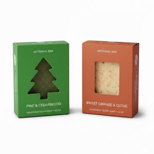 Op de afbeelding: Twee dozen ambachtelijke zeep. Eén is groen met een dennenboomuitsparing, met het opschrift "Pine & Cedarwood". De andere is oranje, met een venster dat de zeep laat zien, met het opschrift "Sweet Orange & Clove". Beide dozen zeggen "Handgemaakte Vegan Zeep - 127.5 g."