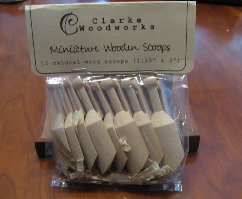 May include: A package of 12 natural wood miniature scoops, each measuring 1.25 inches by 3 inches. The scoops are packaged in a clear plastic bag with a brown paper label that reads "Clarke Woodworks Miniature Wooden Scoops".
