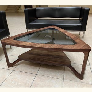 Puede incluir: Mesa de centro triangular con estructura de madera oscura y tapa de cristal. La mesa tiene un estante inferior y patas anguladas. Sus dimensiones son de aproximadamente 100 cm de ancho y 90 cm de profundidad.