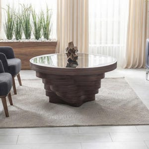 May include: A modern living room arrangement with a dark wood coffee table (approx. 100 cm diameter) with a glass top, two grey armchairs, and a navy blue sofa. The coffee table has a layered design. A neutral rug and plants.