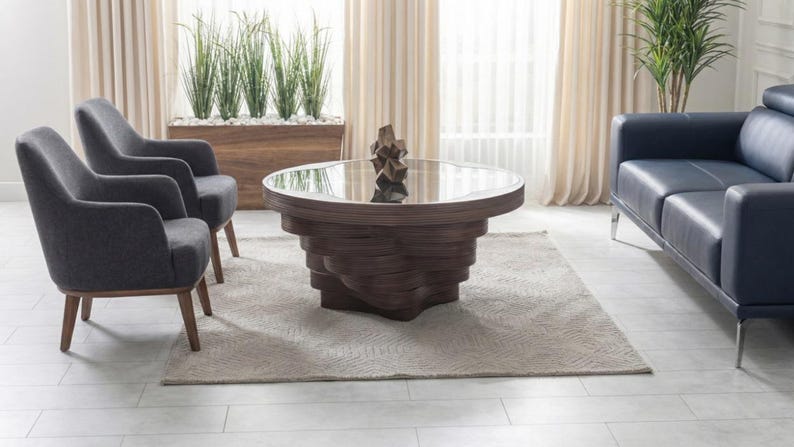 May include: A modern living room arrangement with a dark wood coffee table (approx. 100 cm diameter) with a glass top, two grey armchairs, and a navy blue sofa. The coffee table has a layered design. A neutral rug and plants.