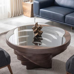May include: A contemporary coffee table with a glass top and a distinctive, swirling wooden base. A bronze sculpture sits on the table. Two grey armchairs and a blue sofa are in the background.