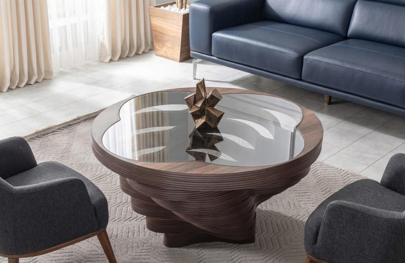 May include: A contemporary coffee table with a glass top and a distinctive, swirling wooden base. A bronze sculpture sits on the table. Two grey armchairs and a blue sofa are in the background.
