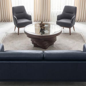 May include: A contemporary living room arrangement with a navy blue leather sofa, two grey armchairs, and a round coffee table with a glass top. The coffee table has a distinctive wooden base. The room is decorated in neutral tones and natural light.
