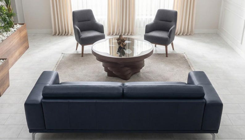 May include: A contemporary living room arrangement with a navy blue leather sofa, two grey armchairs, and a round coffee table with a glass top. The coffee table has a distinctive wooden base. The room is decorated in neutral tones and natural light.