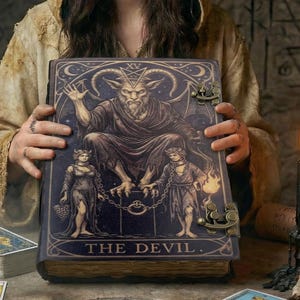 Könnte beinhalten: Eine Person hält ein großes, verziertes Buch mit dem Titel "THE DEVIL". Das Buch zeigt eine detaillierte Illustration einer gehörnten Figur und zwei gefesselten Figuren. Tarotkarten, Kerzen und eine Feder befinden sich auf dem Tisch. Die Szene hat ein mystisches, gotisches Thema.