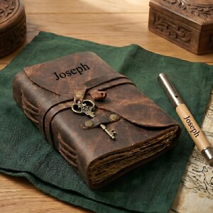 May include: A brown leather journal with the name "Joseph" embossed on the cover, secured with a strap and key clasp. A matching pen with "Joseph" printed on it rests nearby. The journal is on a green cloth, with wooden boxes in the background.