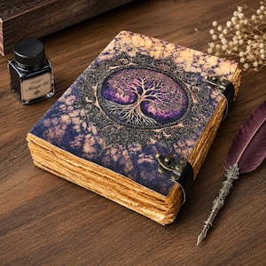 May include: A rectangular journal with a purple and gold marbled cover featuring a central tree illustration inside an ornate circular frame. The book has thick, deckled-edge pages, two black leather straps with brass metal clasps, a bottle of ink, and a quill.