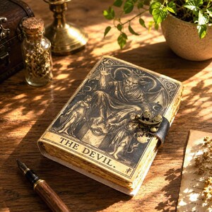 May include: A rectangular journal with a cream-coloured cover featuring a black ink illustration of The Devil tarot card, including a horned figure, two chained individuals, and the text THE DEVIL. The book has a dark metal latch closure.
