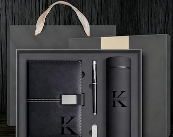 May include: A black gift set featuring a leather-bound notebook, pen, and tumbler, all personalized with the name "Kenneth" and the initial "K". The set is presented in a gray box with a matching gift bag.